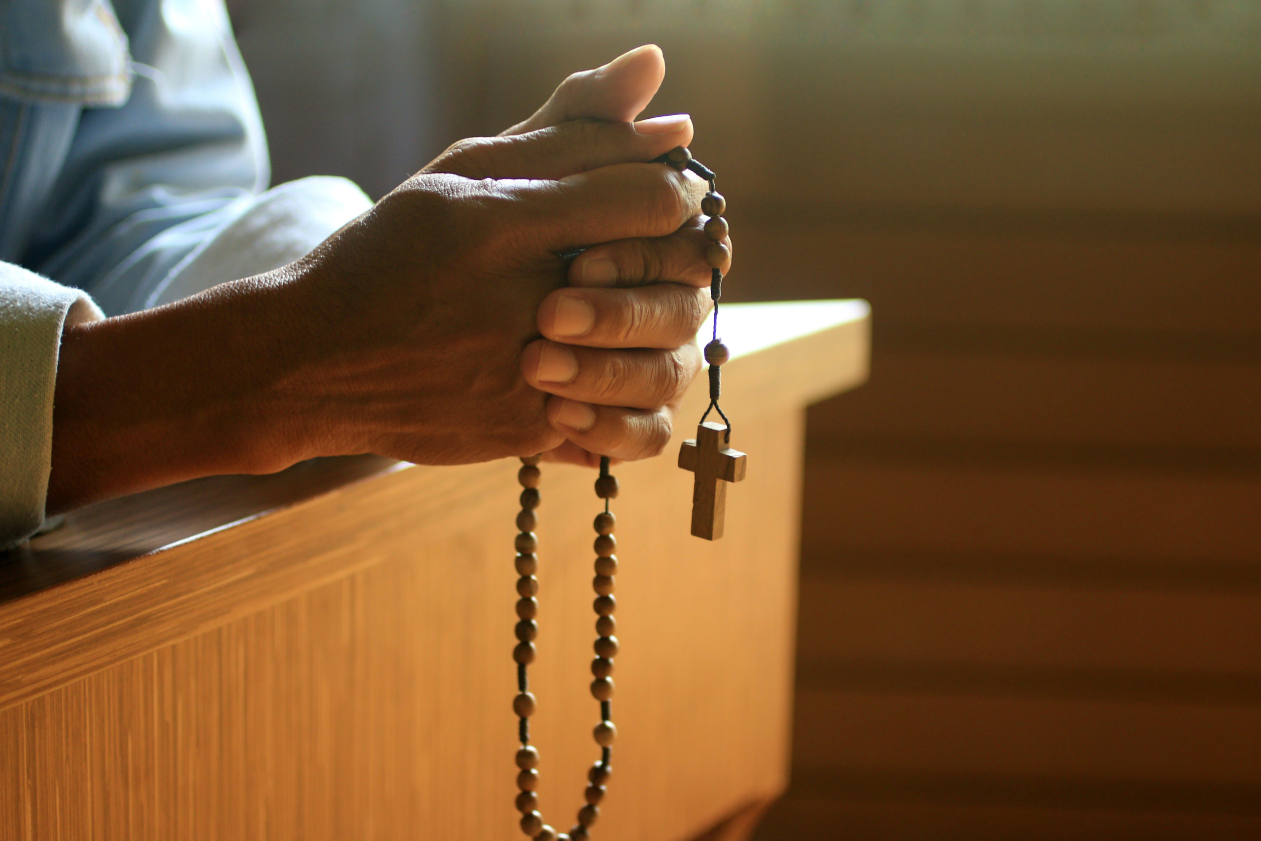 Senior man kneel, holding wooden rosary beads in hand with Jesus Christ holy cross crucifix in the church. Natural light background. Prayer pose crop closeup with copy space. Catholic symbol of faith.