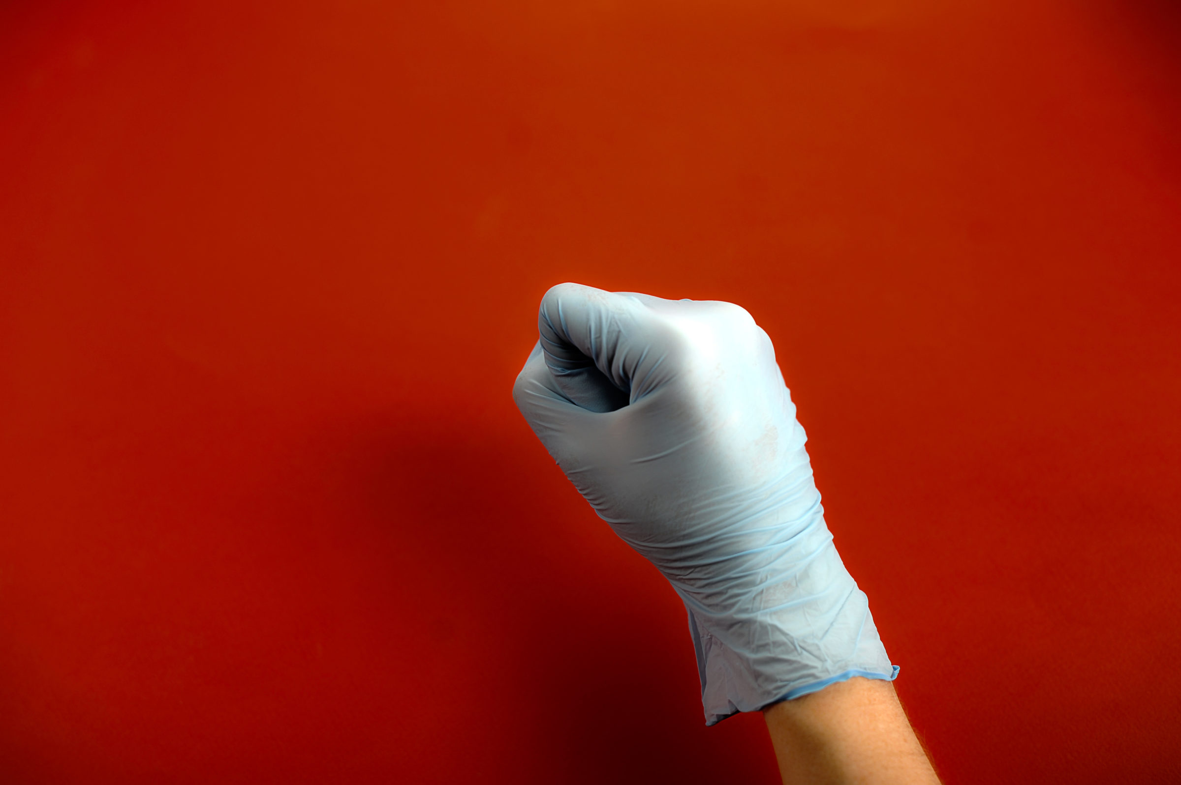Hand in medical latex glove. Hand clenched into a fist. Red background. Close-up. Concept: Hand gestures for expressing emotions.