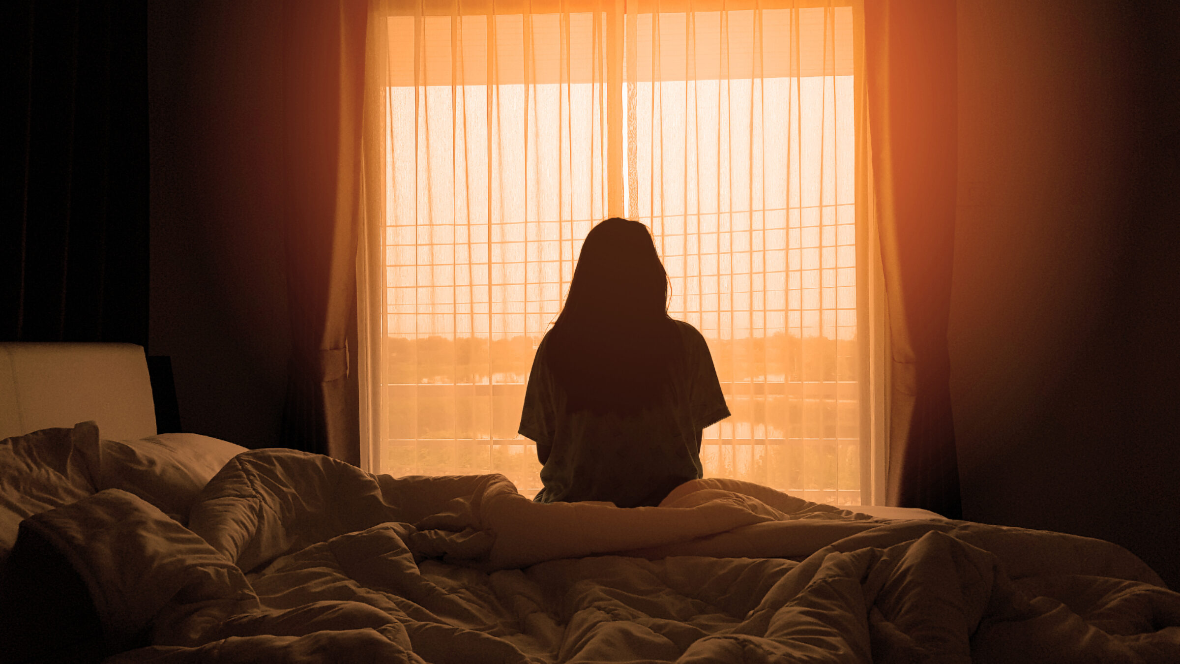 silhouette of woman sitting on the bed beside the windows with sunlight in the morning